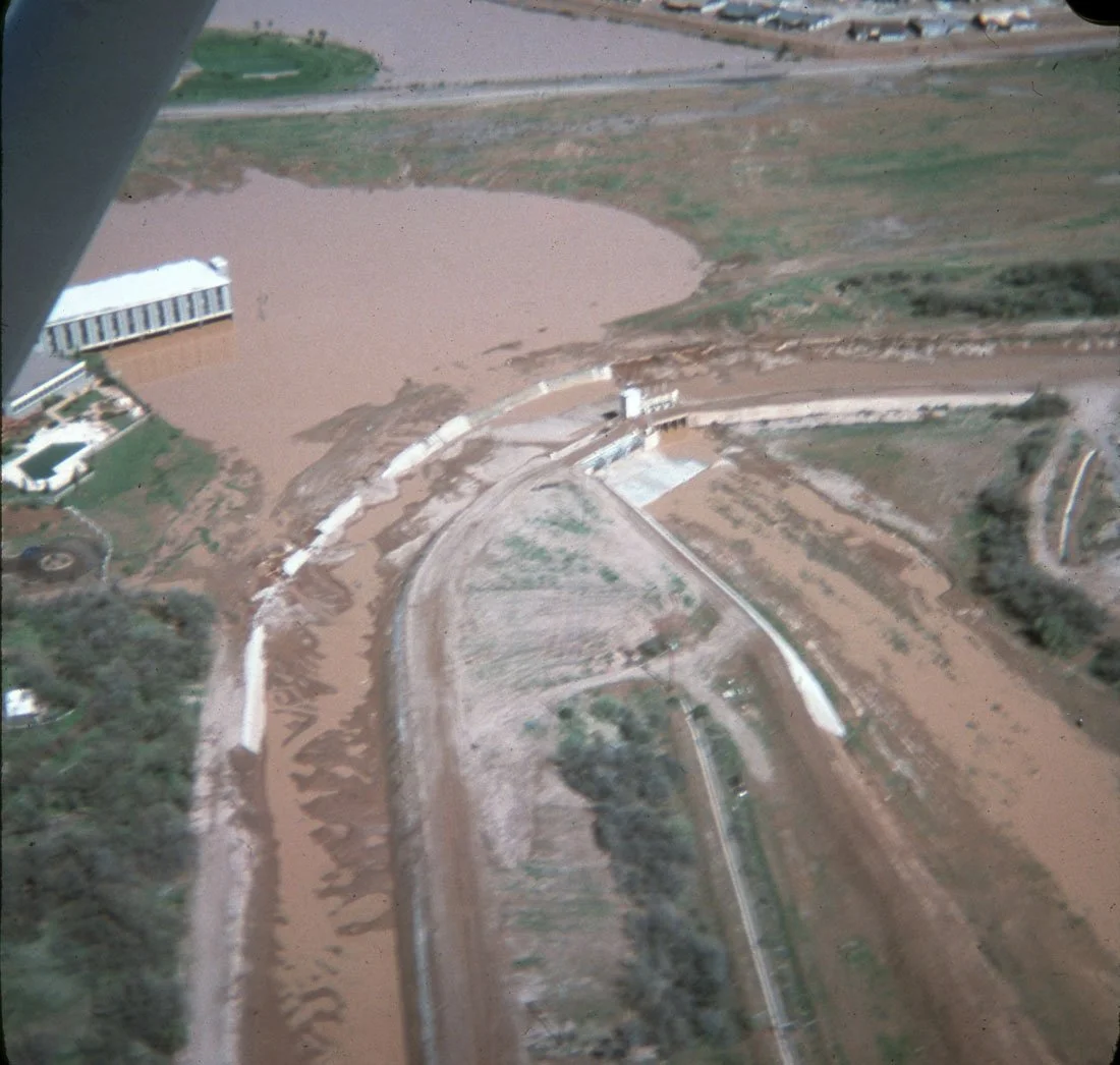 An aerial view of farmland with tilled soil, some green patches, and buildings, possibly from a crop duster or other aircraft.