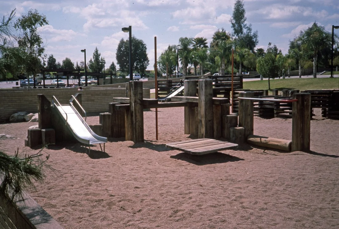 Wooden playground structure with slides, a bridge, and climbing elements on a sandy surface in a park.
