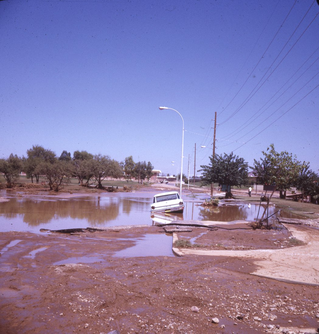Flooded street with a buried car and water covering the road, trees, streetlights, and power lines.