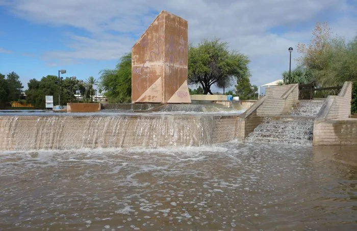 A fountain with water cascading over a ledge, featuring a large vertical sculpture and surrounding trees and stairs.