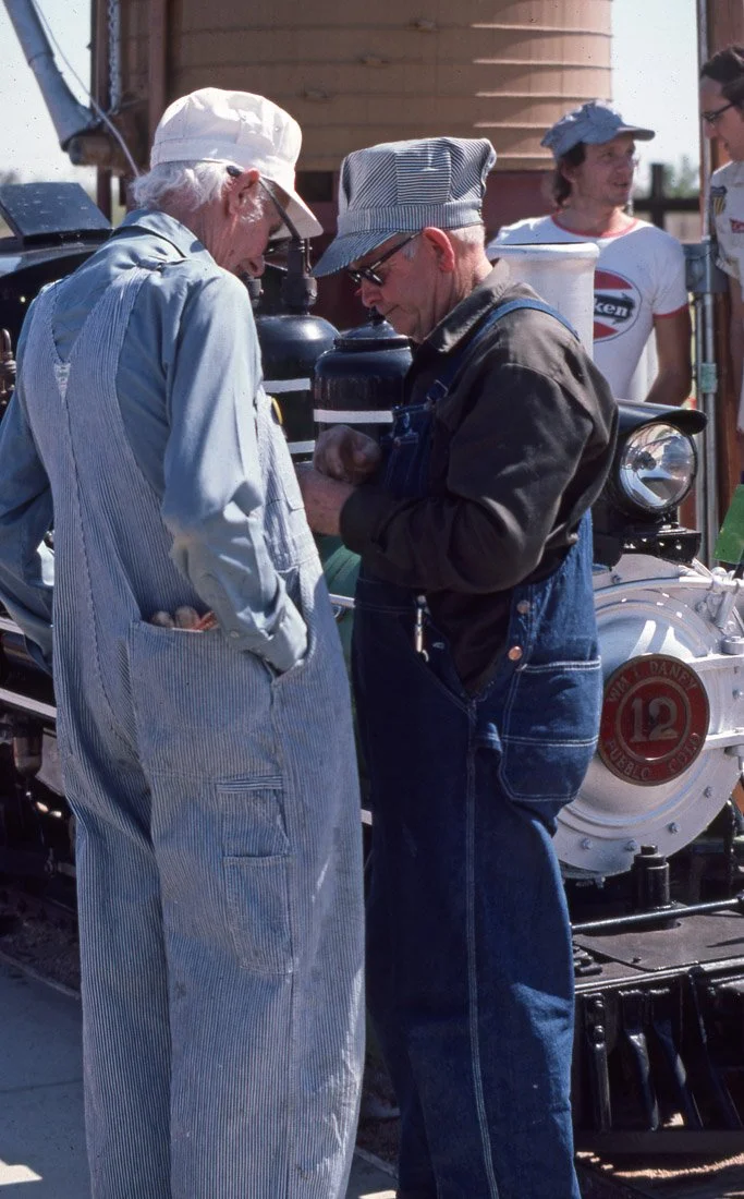 Two elderly men wearing work clothes and hats, engaged in conversation next to a vintage train engine with the number 12, while two younger women stand in the background near a water tower.