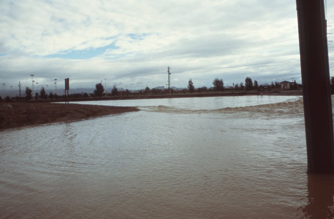Flooded area with water covering the ground, cloudy sky, utility poles, and some distant trees and buildings.