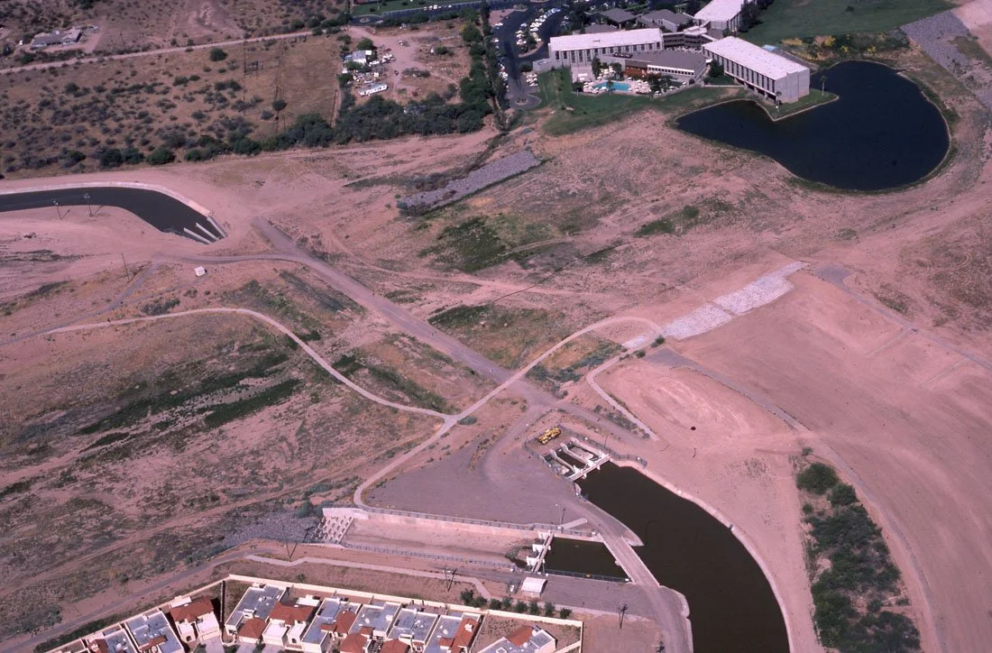 Aerial view of a dam and water reservoir in a semi-arid landscape, with surrounding roads and a few buildings