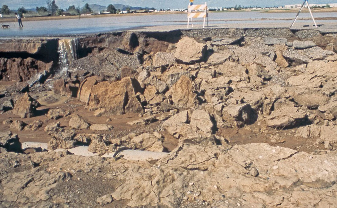 View of a large area with broken asphalt and rocks, possibly a road or pavement under construction or repair, with a road on top and orange safety barriers in the background.