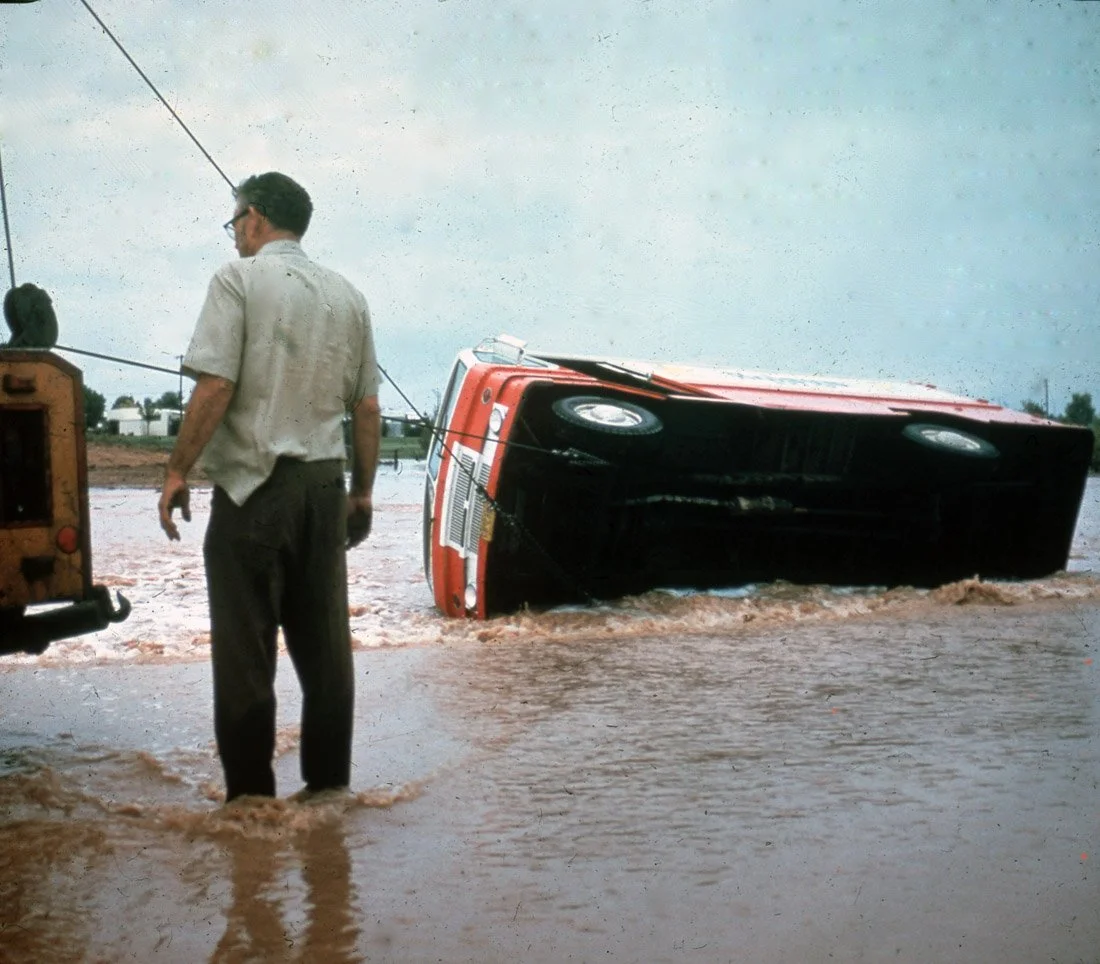 A red vehicle has flipped onto its side in floodwaters during a storm, with a man standing nearby looking at the scene.
