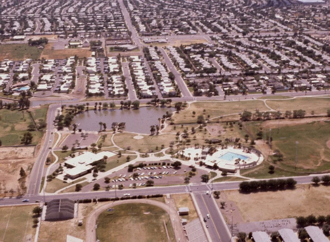 Aerial view of a park with a lake, swimming pool, and parking lot, surrounded by roads and residential neighborhoods.