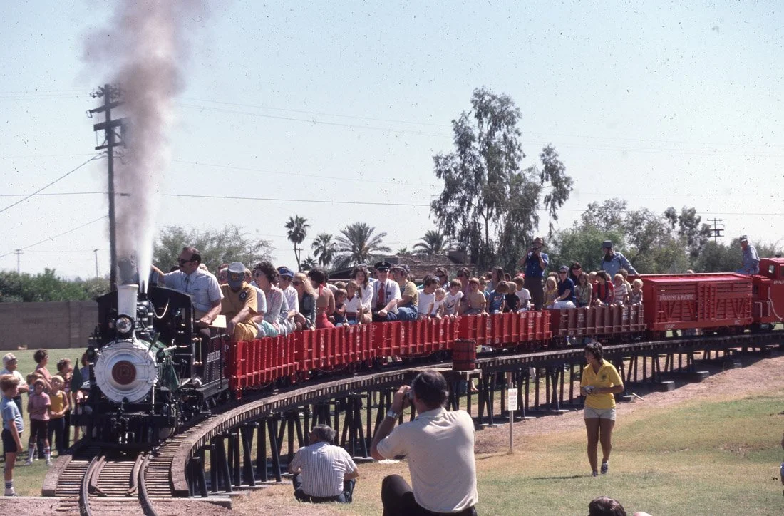 People riding a red open-air train through a park with trees and palm trees in the background, while others watch or take photos.