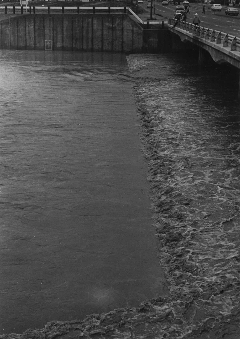 Black and white photo of water flowing beneath a bridge with a city street above, showing cars and pedestrians.