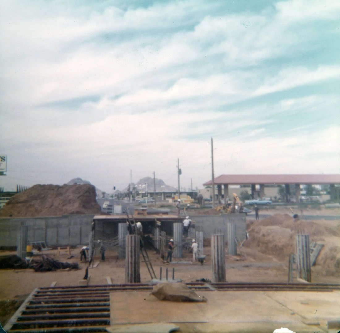 Construction site with workers assembling concrete forms and building a structure. Dirt piles and construction equipment are visible, with a partly cloudy sky overhead.