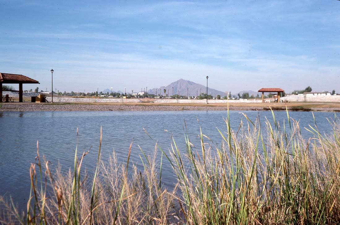 A body of water with tall grass in the foreground, a paved area with lamp posts, gazebos, and a mountain in the background.