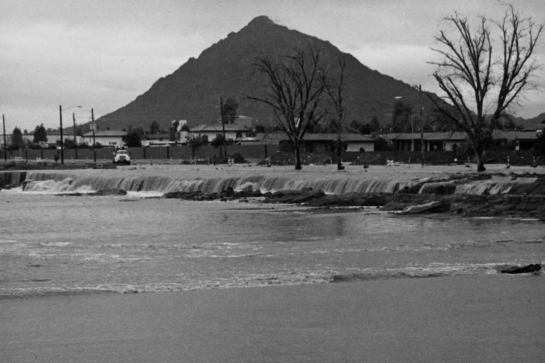 Black and white photo of a river with a small waterfall, trees without leaves on the riverbank, and a mountain in the background.