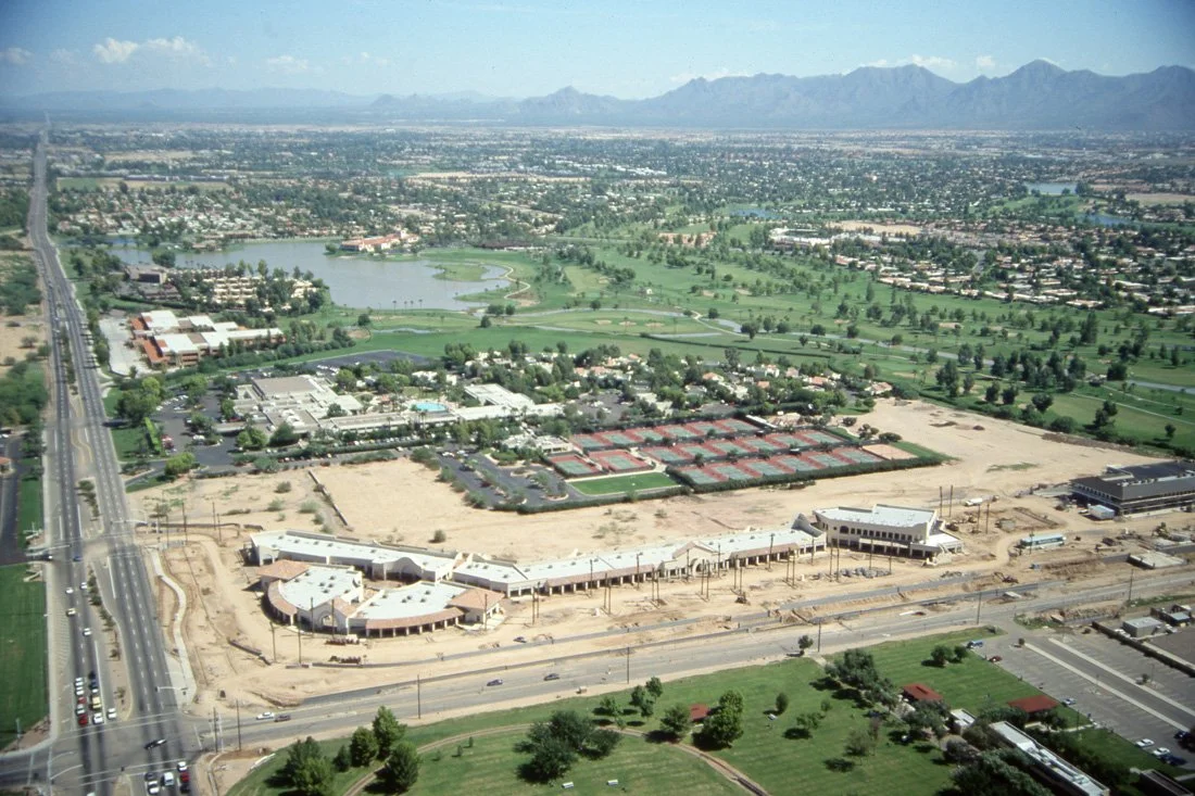 Aerial view of a city with a golf course, a lake, and multiple buildings, including a construction site in the foreground and mountains in the background.