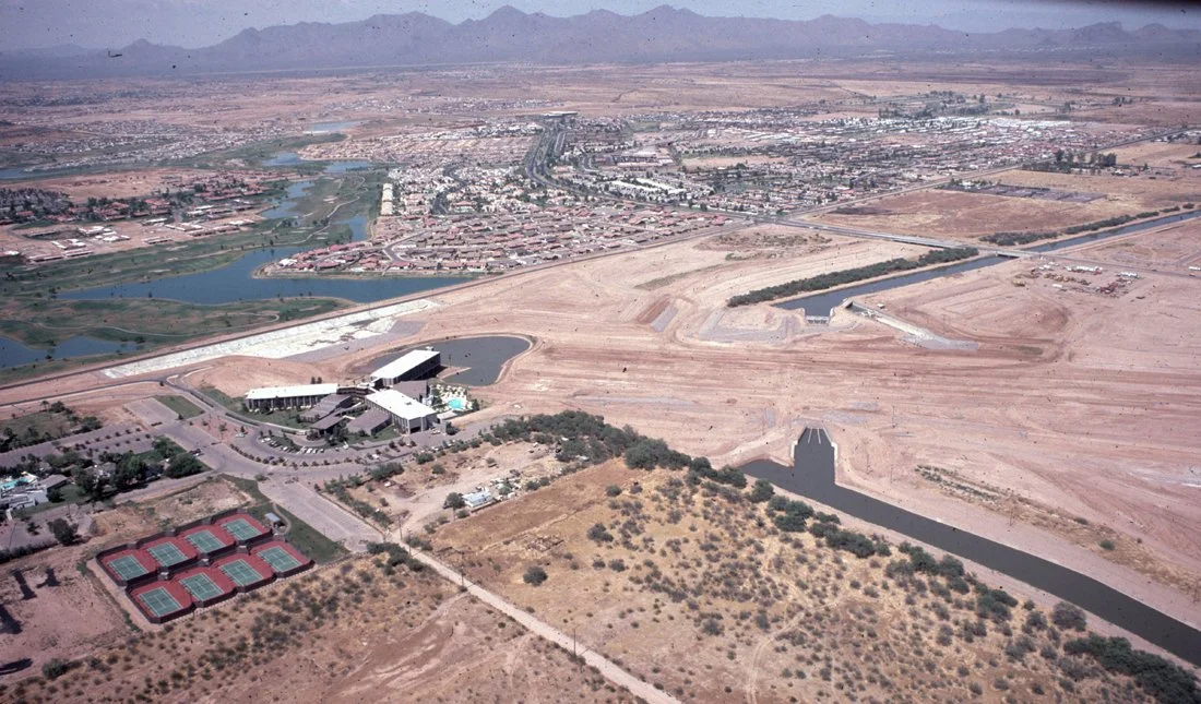 Aerial view of a small town with a river, multiple buildings, and tennis courts in a desert landscape, with mountains in the background.