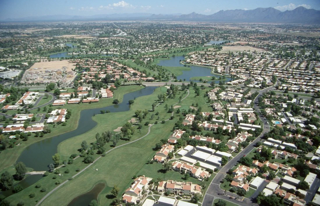 Aerial view of a suburban city with a golf course and water features, residential neighborhoods, and distant mountains under a blue sky.