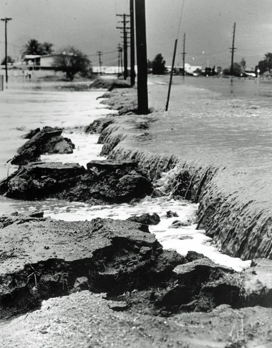 Flooded street with water covering the road, damaged pavement, and utility poles leaning with power lines.