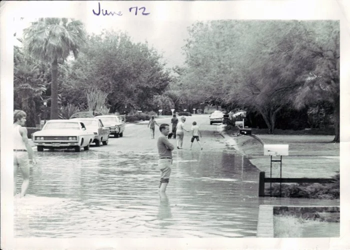 People wading in flooded street lined with trees, cars parked along the street, handwritten note 'June 72' at the top.