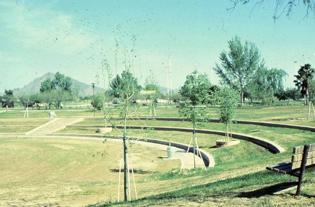 An empty park with small trees, a curved concrete walkway, and a wooden bench on the right. Green grass and scattered trees are visible, with hills in the background.