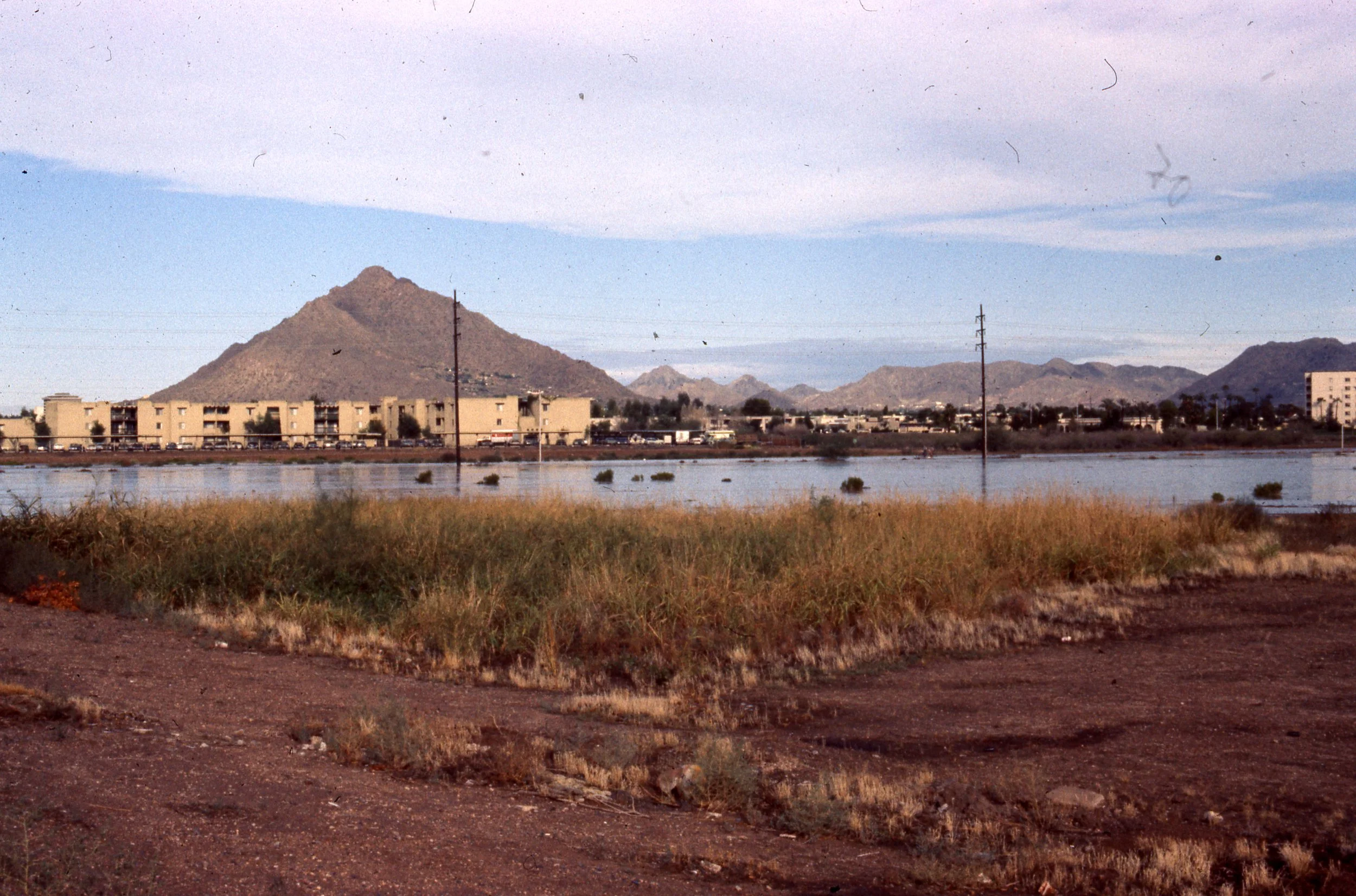 A landscape view featuring mountains in the background, a water body in the midground, and desert-like terrain in the foreground with some grass and shrubs.
