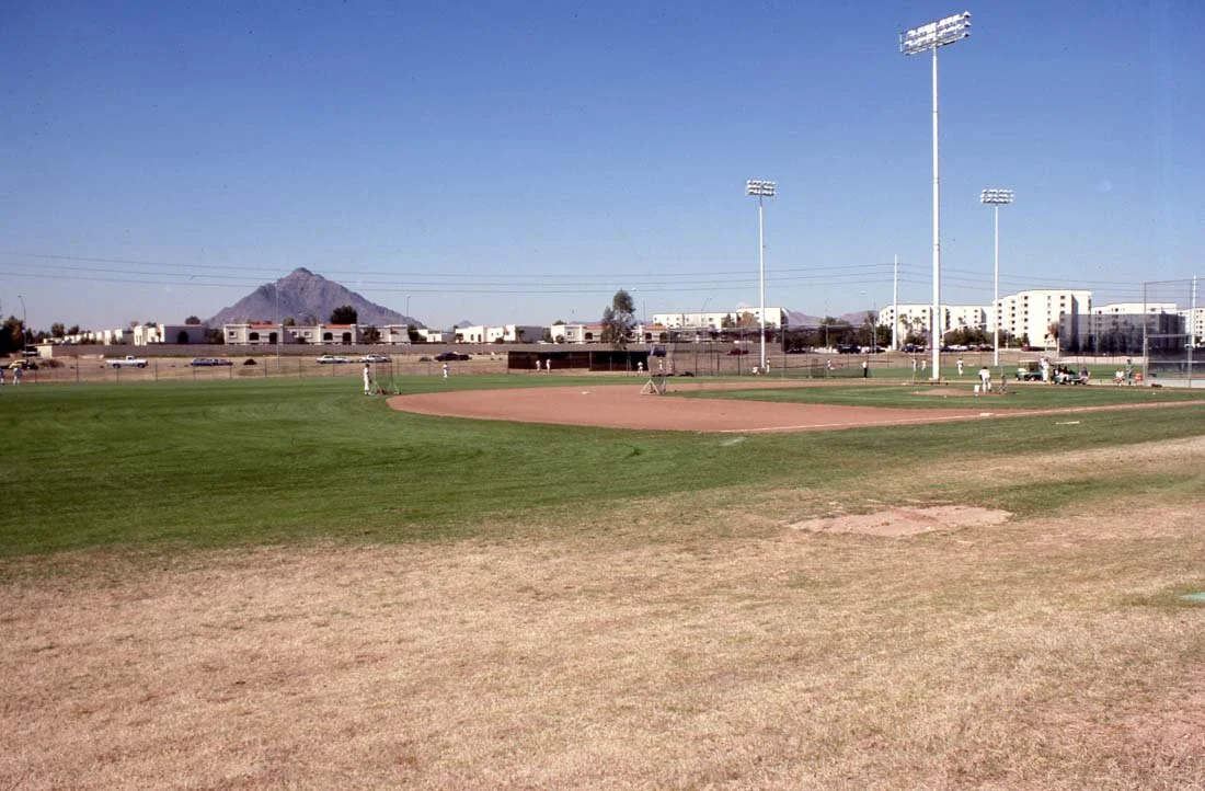 A baseball field with players practicing, surrounded by a grass field, with training equipment and floodlights. In the background, there are several white buildings and a mountain under a clear blue sky.
