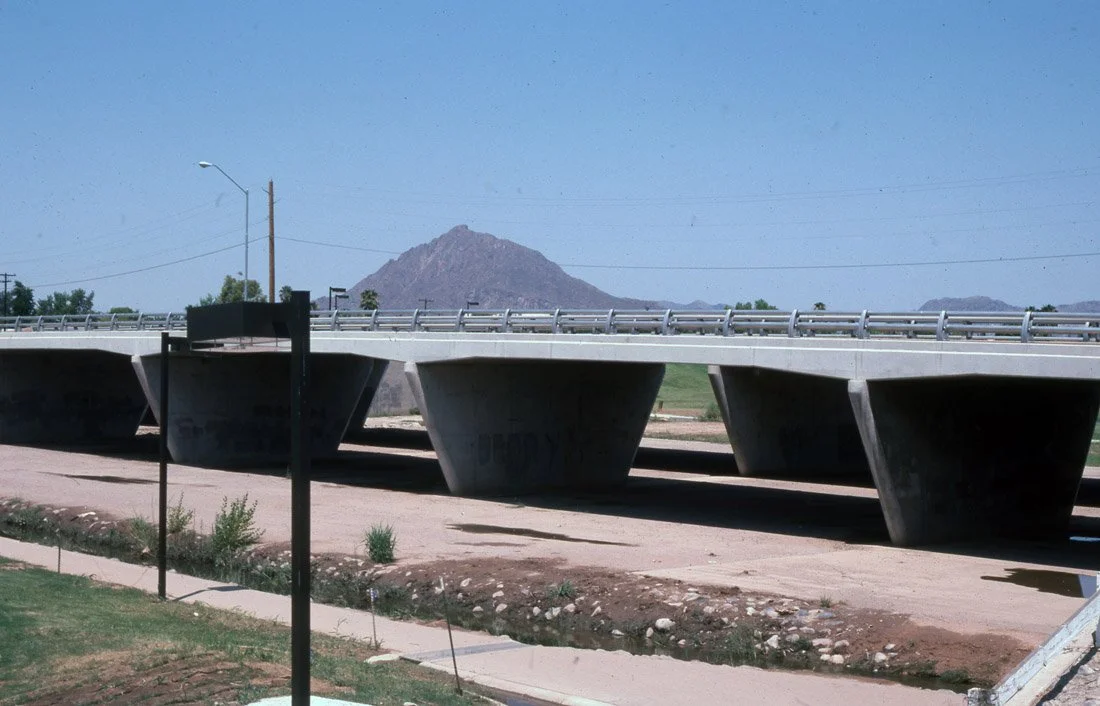 A concrete bridge with large supporting piers over a dirt area, with a mountain in the background and a street lamp on the side.