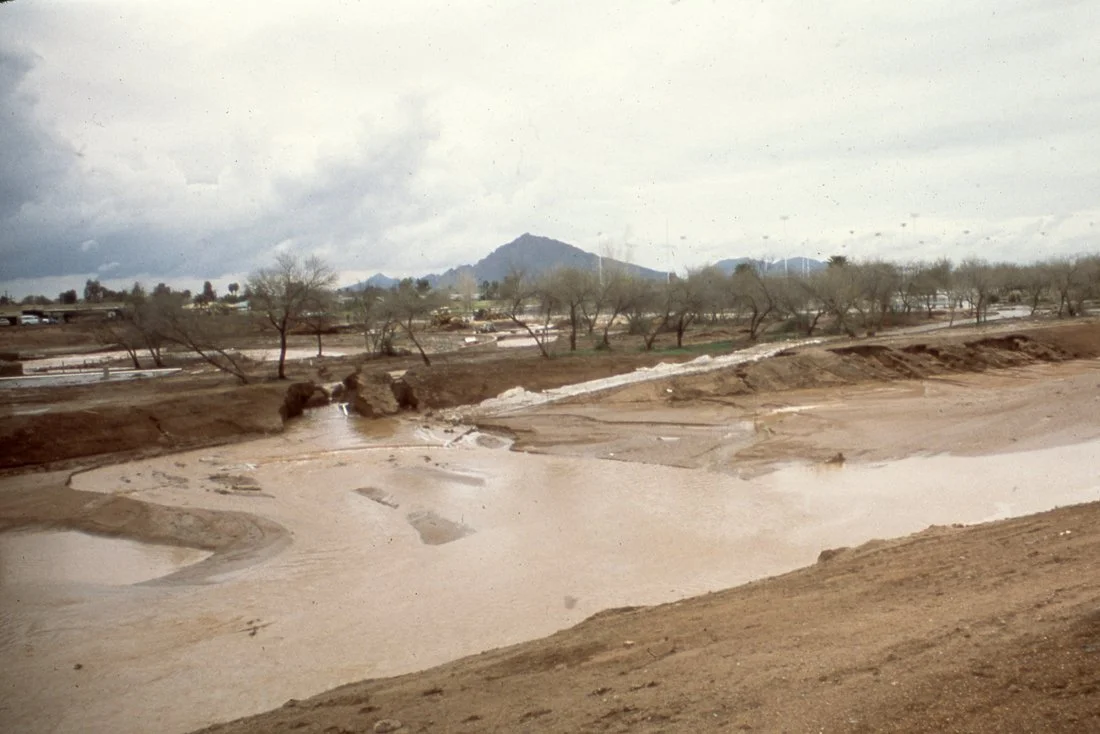 A dry, muddy area with water flowing through it, surrounded by leafless trees and distant mountains under a cloudy sky.