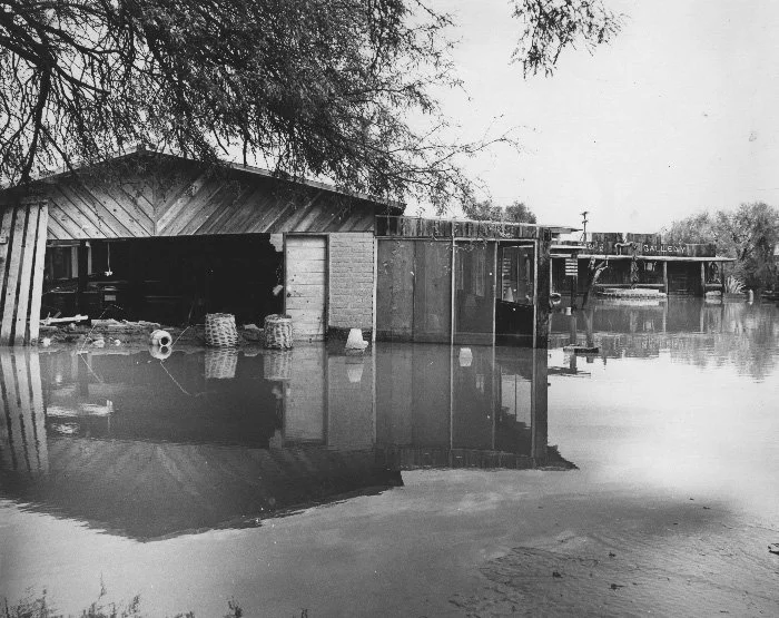A flooded area with a partially submerged wooden building and a boat docked nearby, surrounded by water and trees.