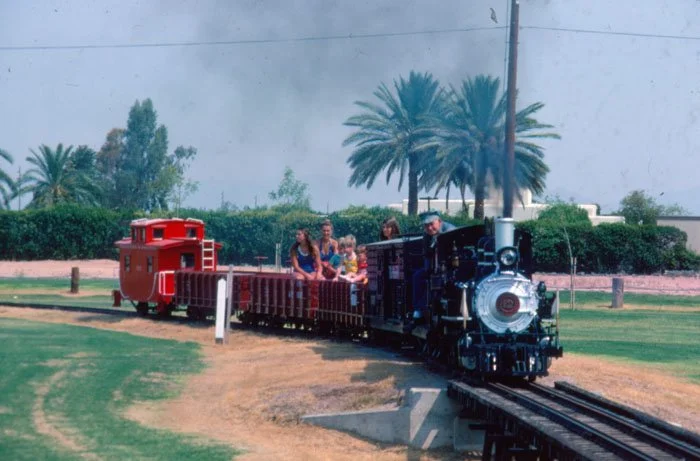 Child and adult riding on a miniature train in a park with palm trees in the background.
