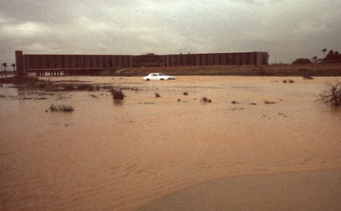 Flooded desert landscape with a white car and a large building in the background.