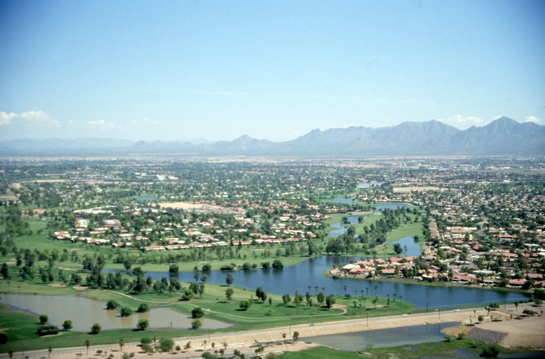 Aerial view of a city with lakes, green spaces, residential neighborhoods, and mountains in the background under a clear blue sky.