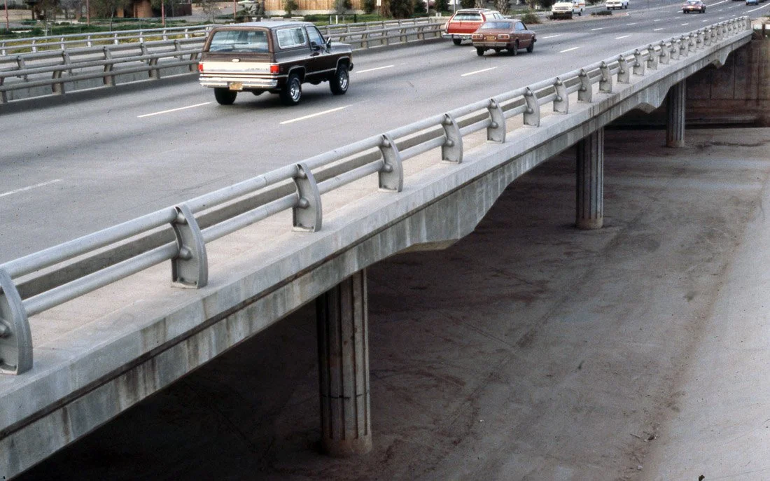 A concrete highway overpass with metal guardrails, passing over an empty dirt area below, with several cars driving on the road.