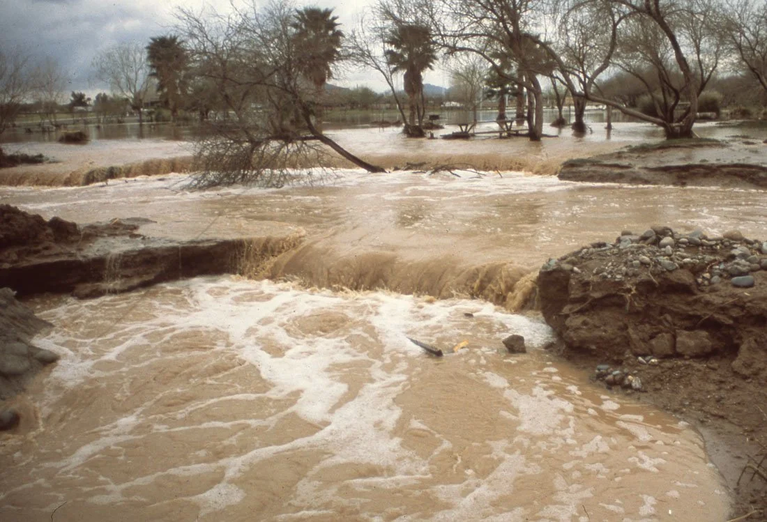 A flooded area with muddy water flowing over a small waterfall, surrounded by trees and rocks.