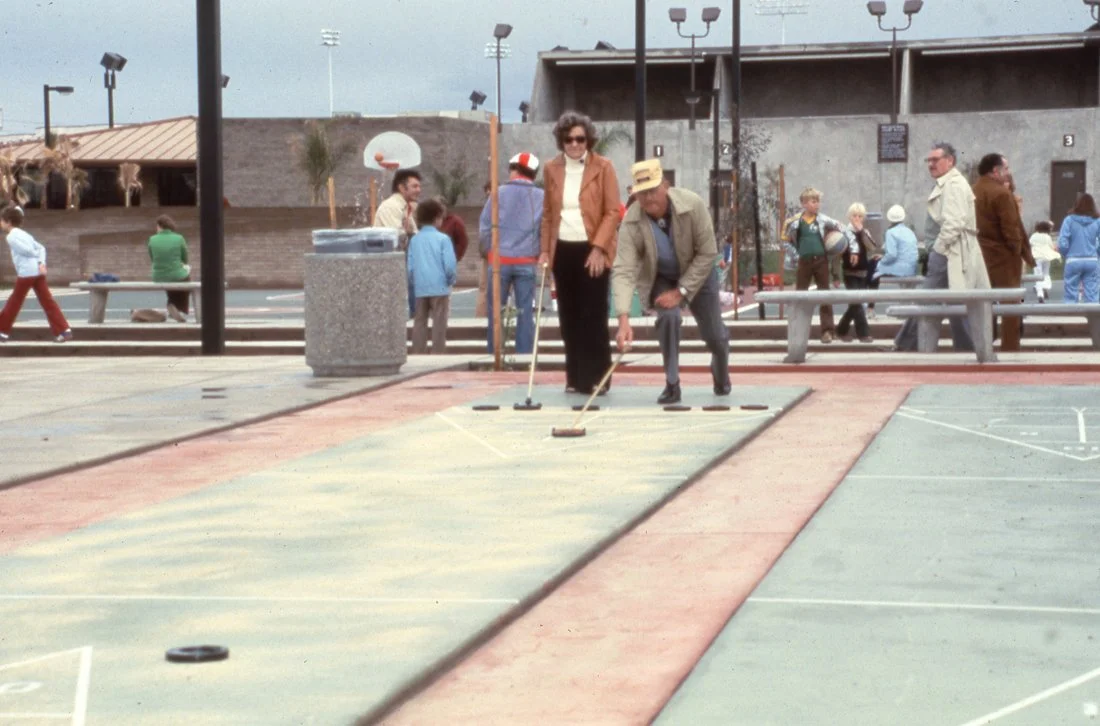 People playing shuffleboard on an outdoor court surrounded by spectators, with a basketball hoop in the background.