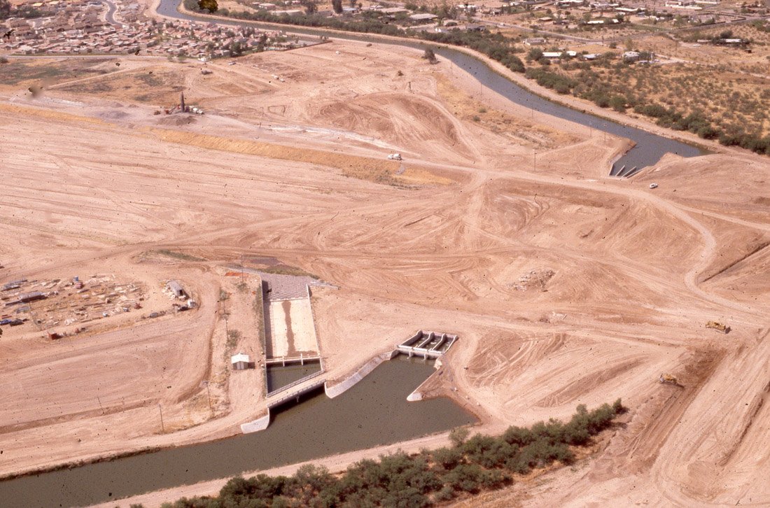 Aerial view of a dam under construction in a dry area, with a river flowing through it and surrounding dirt roads and land.