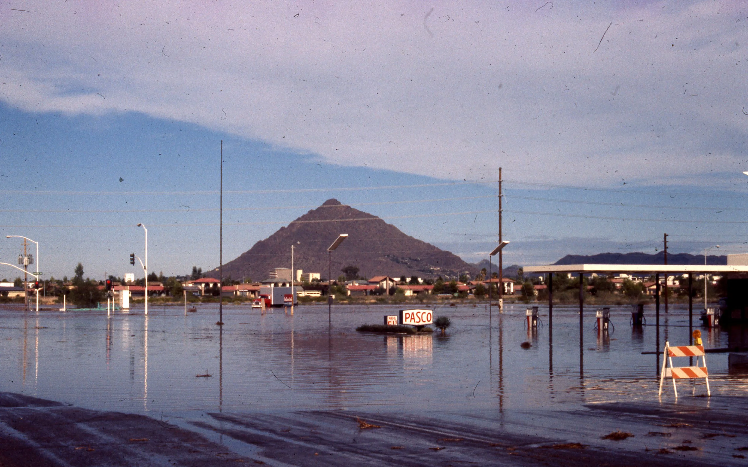A parking lot that is flooded with water, with submerged signs and lampposts in the foreground. In the background, there is a mountain and a mostly cloudy sky.