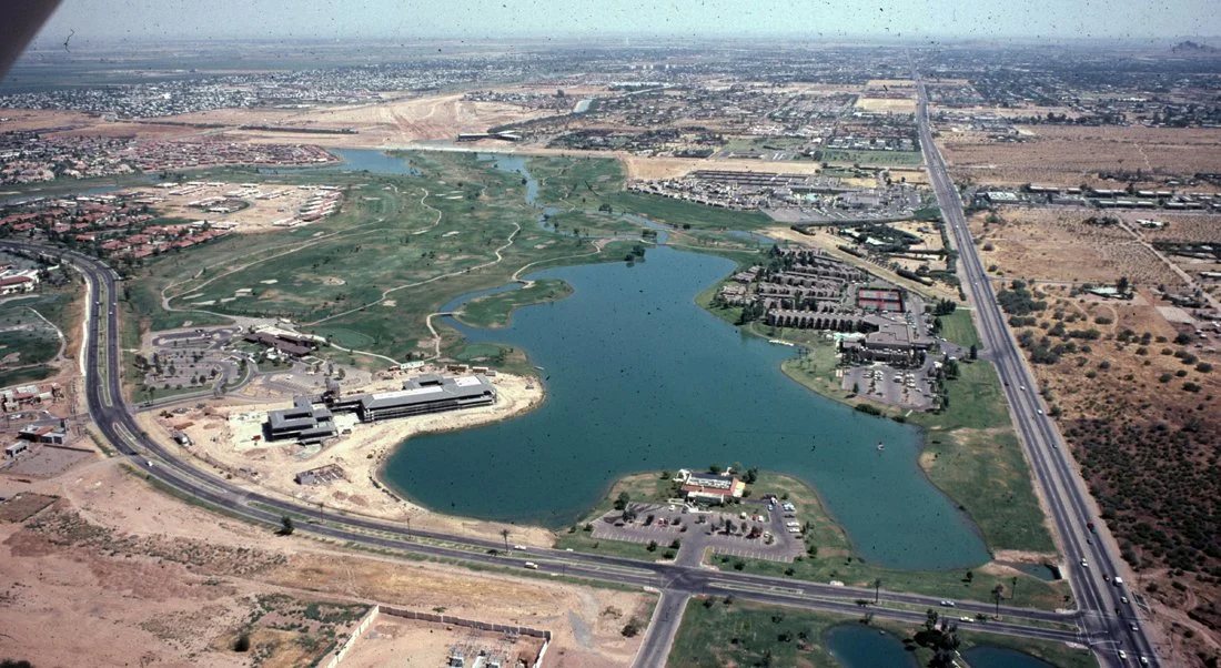 Aerial view of a large lake surrounded by roads, buildings, and open land, with a cityscape in the distance.
