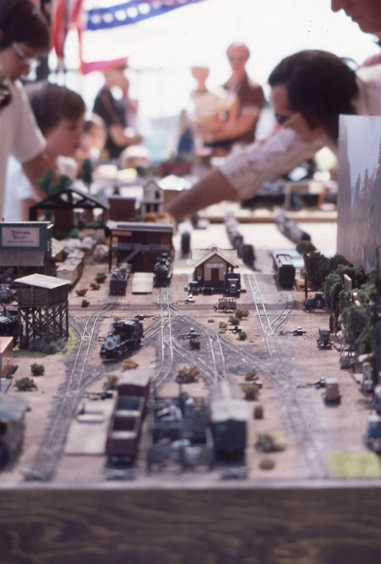 A model train setup with tracks, trains, and miniature buildings, surrounded by people, some of whom are pointing at or observing the display.