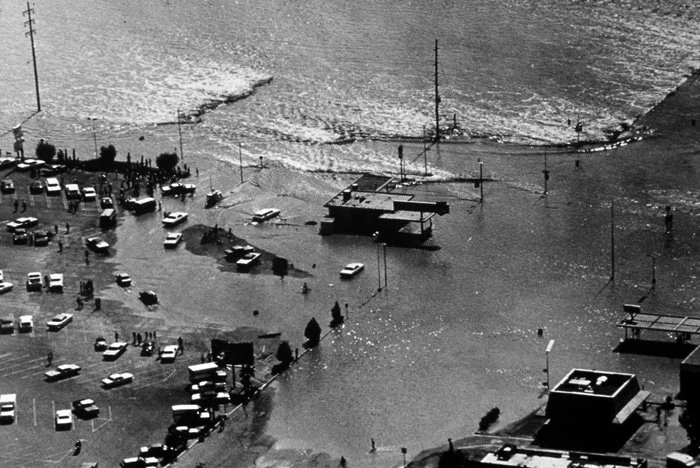A flood has submerged a parking lot and part of a street near the coast, with water spreading over parked cars and streets, and waves visible in the background.