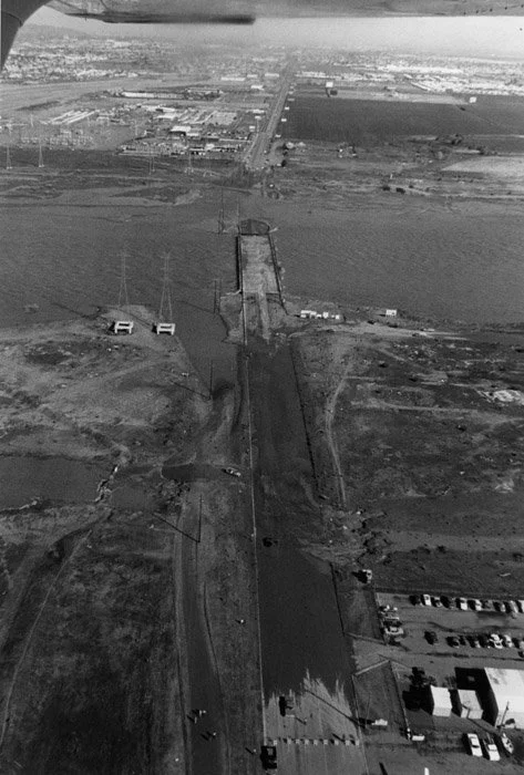 Aerial view of a dam constructed across a river with spillway, with surrounding land, utility poles, and buildings in the distance.