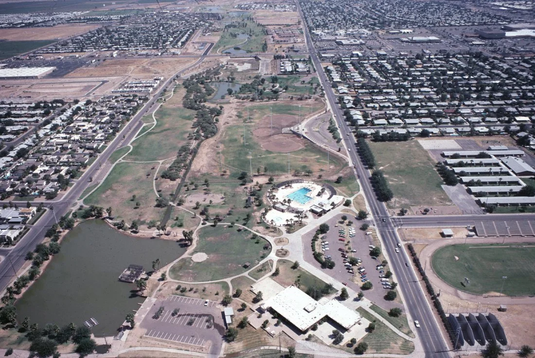 Aerial view of a park with a pond, walking paths, a swimming pool, sports fields, and surrounding city streets and neighborhoods.
