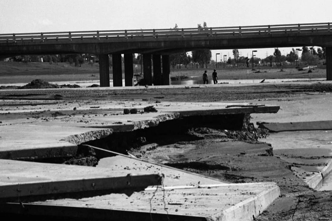 Damaged pavement with cracked and broken asphalt and concrete, and a bridge over a river in the background.