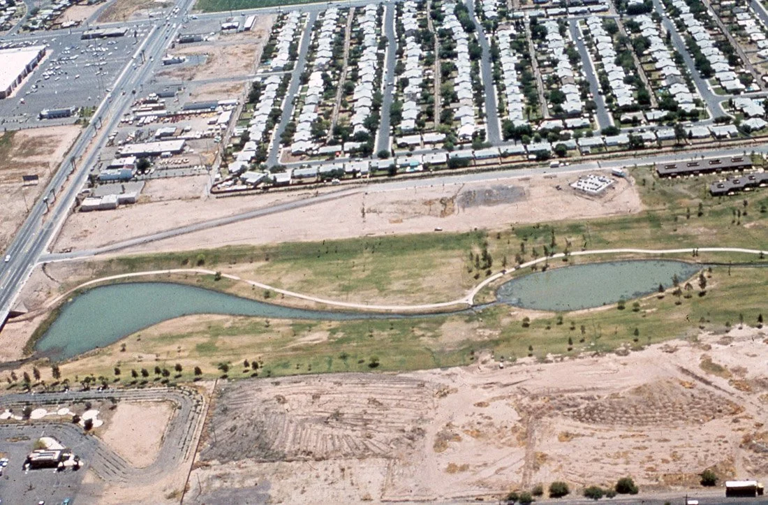 Aerial view of a park with two shaped ponds, grass, trees, and walking paths, adjacent to a residential area with houses, a commercial area, and a highway.