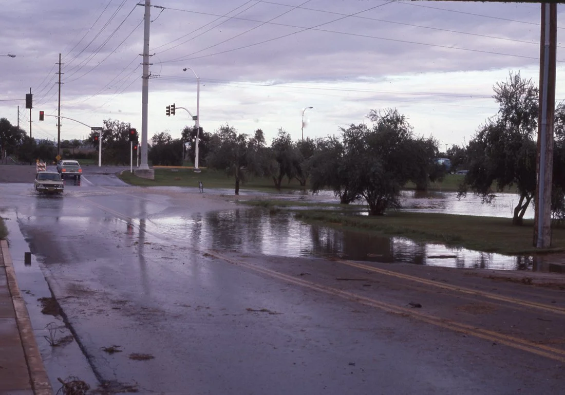 A flooded street with water covering the road, trees, utility poles, and a traffic light, with cloudy skies overhead.