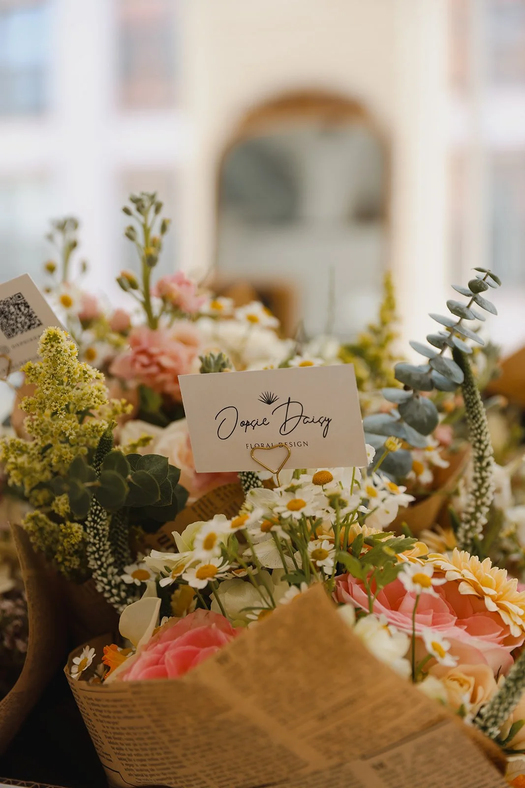 A bouquet of assorted flowers with a business card reading 'Topsie Daisy Floral Design' on top, placed on a surface with a blurred mirror or window in the background.