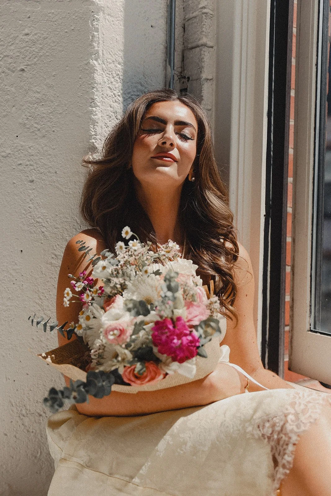 A woman with long brown hair sitting by a window, holding a bouquet of pink, white, and purple flowers with greenery, wearing a cream-colored dress with lace details.