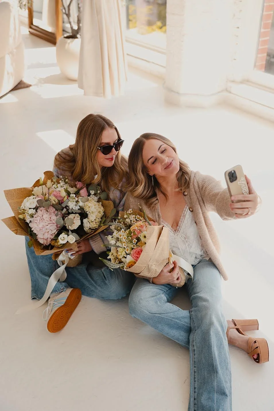 Two women sitting on the floor, taking a selfie. They are holding bouquets of flowers and smiling. One woman is wearing sunglasses, the other has her eyes closed. Bright, sunlit room with large windows in the background.