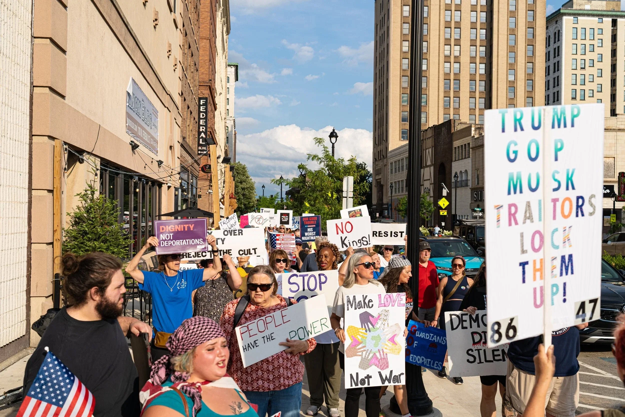 A large group of people participating in a protest march on a city street, holding various signs with messages supporting Dignity, love, and resistance, with tall buildings and a blue sky in the background.