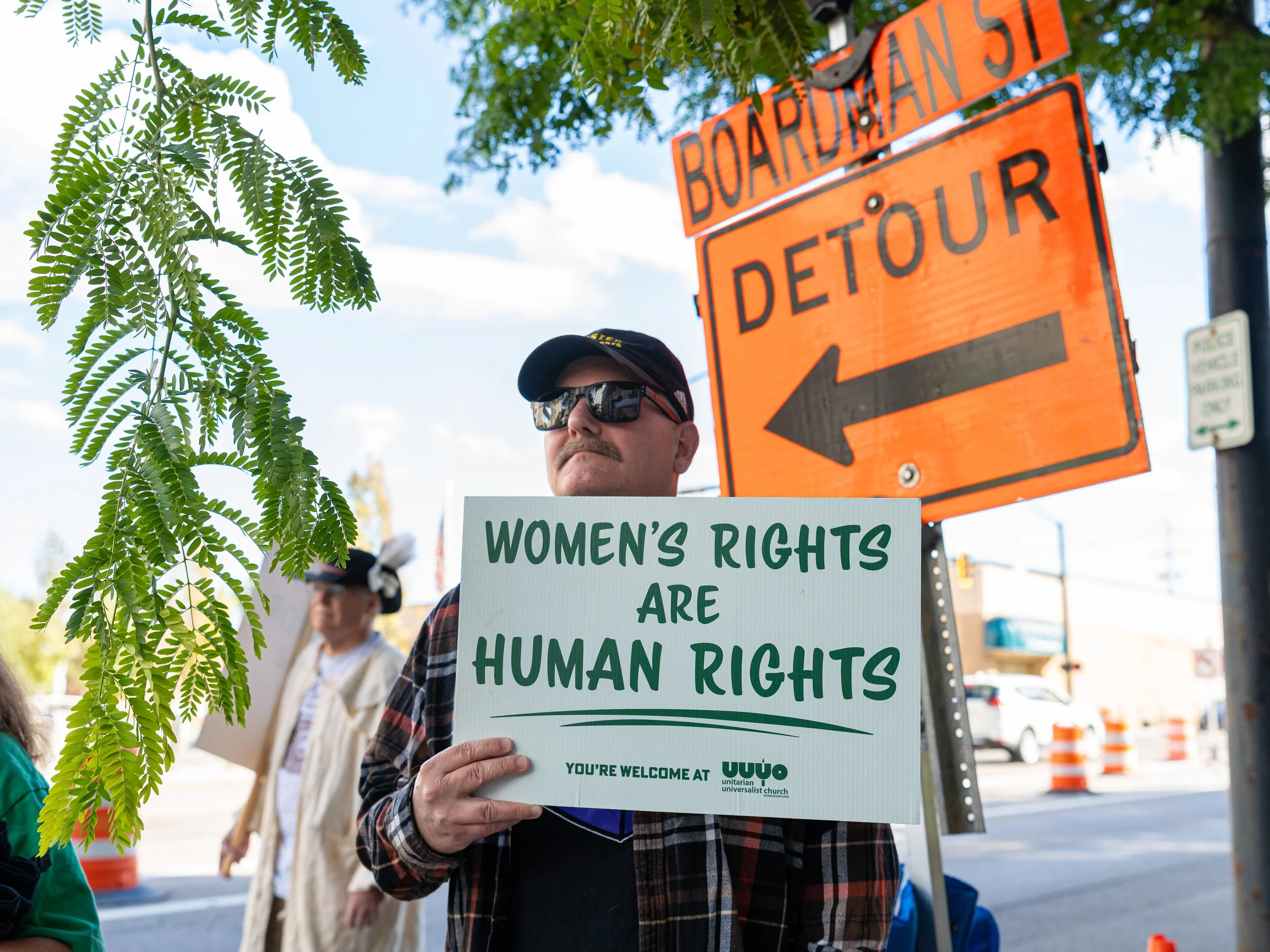 A man holding a sign that reads 'Women's Rights Are Human Rights' participates in a protest or demonstration. The scene takes place outdoors with trees, a street, and a traffic detour sign nearby.