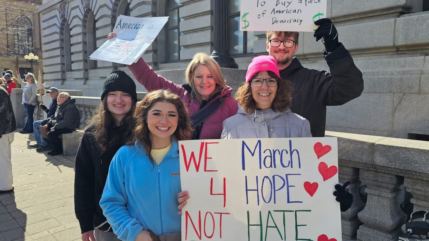 Group of five smiling people participating in a protest or rally, holding signs with messages advocating for hope and unity, standing outside a government building on a sunny day.