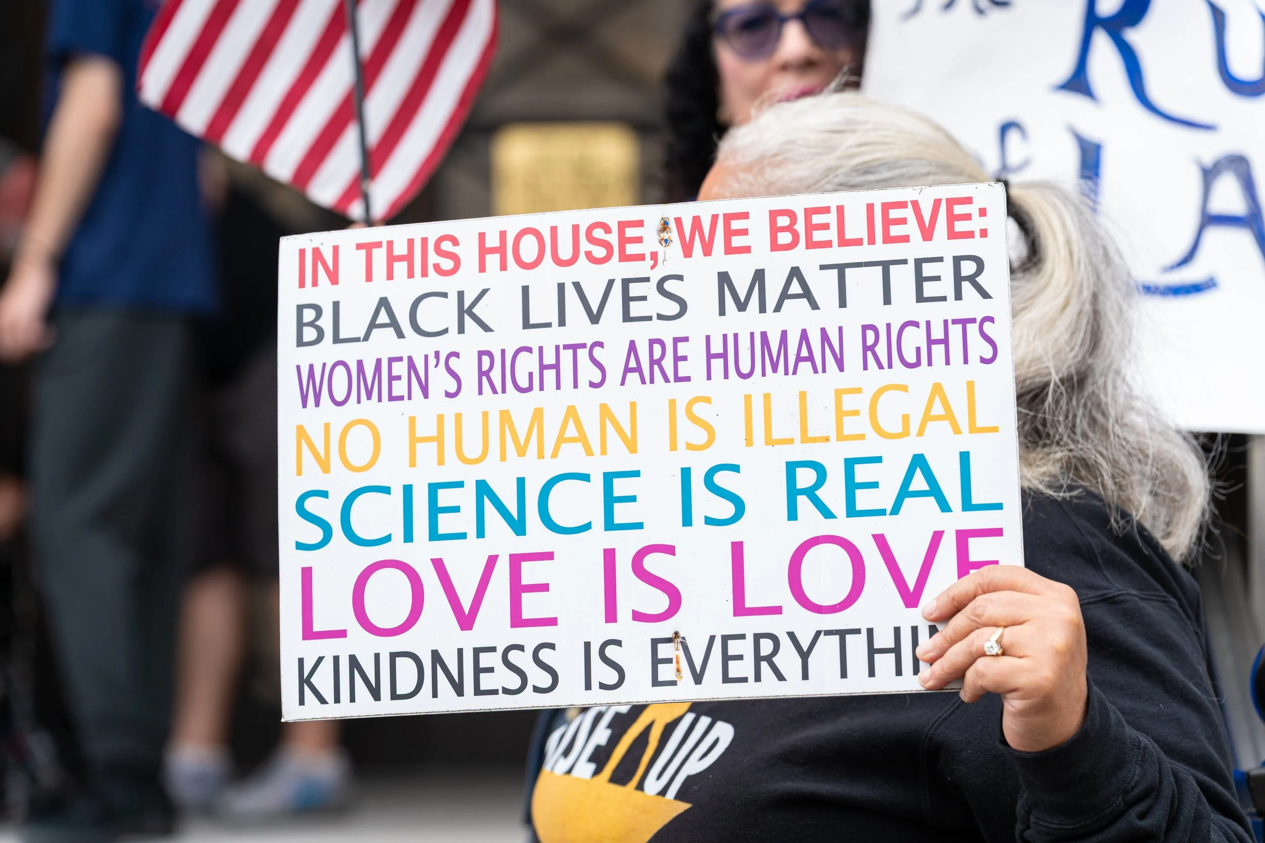 A person holding a sign at a protest that reads, 'In this house, we believe: Black Lives Matter, Women's Rights Are Human Rights, No Human Is Illegal, Science Is Real, Love Is Love, Kindness Is Everything.' In the background, there are other protesters and an American flag.