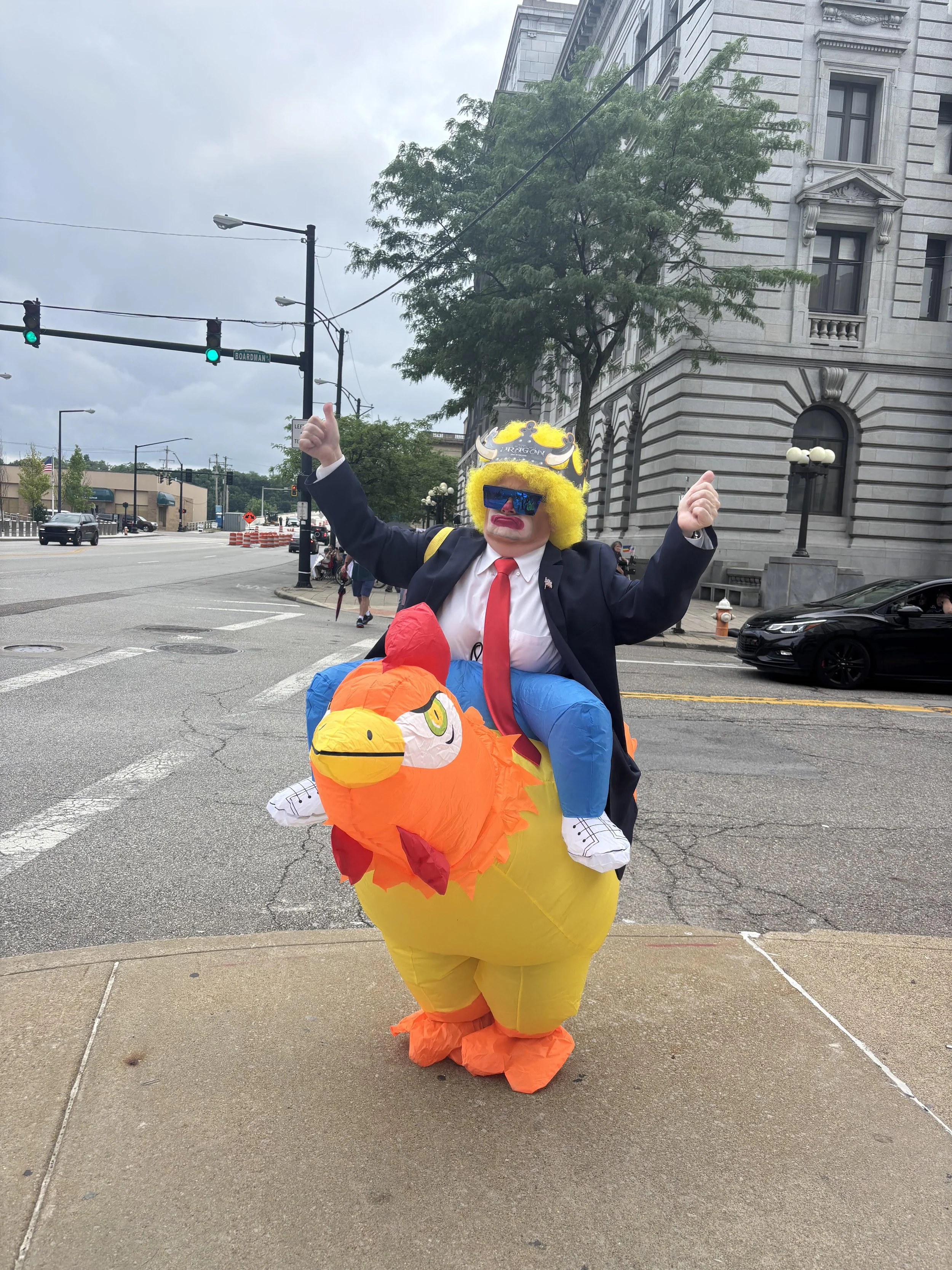 Person dressed as a clown riding a large inflatable chicken costume on city street. Person wears a suit with a red tie, yellow wig, sunglasses, and face paint. The inflatable chicken is orange with a red comb and yellow beak.